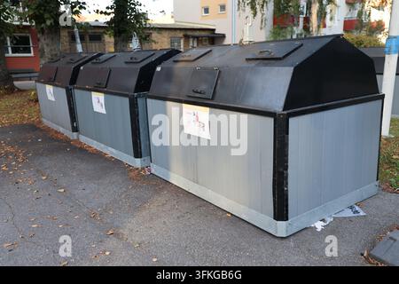 Diversi grandi contenitori di riciclaggio esterni con pannelli di legno grigi e coperchi neri, posizionati sull'asfalto con foglie autunnali vicino a un edificio. Foto Stock