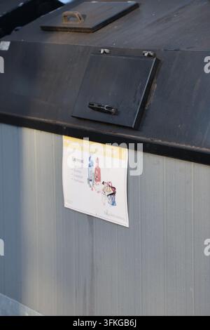 Diversi grandi contenitori di riciclaggio esterni con pannelli di legno grigi e coperchi neri, posizionati sull'asfalto con foglie autunnali vicino a un edificio. Foto Stock