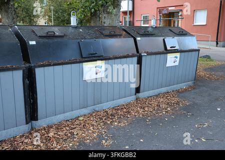 Diversi grandi contenitori di riciclaggio esterni con pannelli di legno grigi e coperchi neri, posizionati sull'asfalto con foglie autunnali vicino a un edificio. Foto Stock