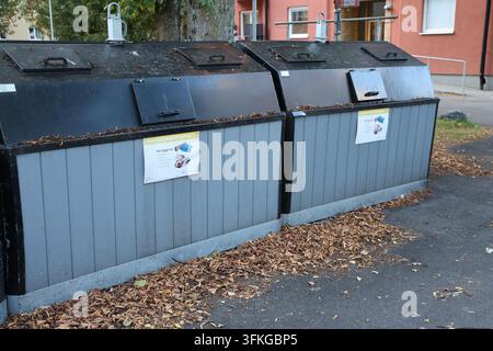 Diversi grandi contenitori di riciclaggio esterni con pannelli di legno grigi e coperchi neri, posizionati sull'asfalto con foglie autunnali vicino a un edificio. Foto Stock