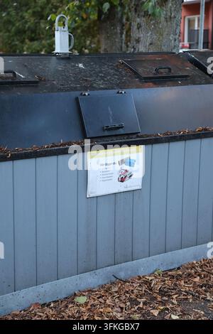 Diversi grandi contenitori di riciclaggio esterni con pannelli di legno grigi e coperchi neri, posizionati sull'asfalto con foglie autunnali vicino a un edificio. Foto Stock