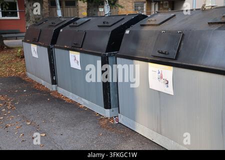 Diversi grandi contenitori di riciclaggio esterni con pannelli di legno grigi e coperchi neri, posizionati sull'asfalto con foglie autunnali vicino a un edificio. Foto Stock