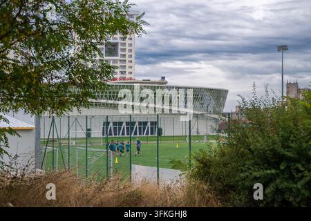 Un'ampia inquadratura vista attraverso alberi e recinzioni che mostra una squadra di calcio giovanile in allenamento su un campo verde davanti a un'arena moderna in vetro curvo e un alto- Foto Stock