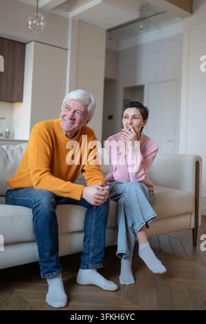 Coppia di anziani felici seduta sul divano a casa, sorridente e che guarda la televisione durante la pensione Foto Stock