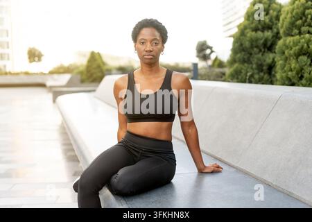 Donna afroamericana seduta su una panchina dopo un allenamento all'aperto indossando abbigliamento sportivo nero. Donna nera che si prende una pausa tranquilla dopo l'allenamento di fitness con un'espressione concentrata. Foto Stock