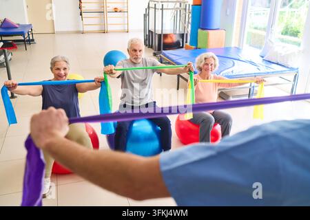 Gli anziani fanno esercizio con bande di resistenza su palline da ginnastica in un centro di riabilitazione supervisionato da un fisioterapista per il recupero e il miglioramento della salute fisica. Questa immagine può essere utilizzata commercialmente per rappresentare la riabilitazione degli anziani nel settore sanitario e Foto Stock