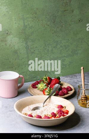 Budino di riso dolce in piatto di ceramica con bacche fragola e lampone, noci, miele e tazza di latte su tavolo da cucina grigio con parete verde come sfondo. Foto Stock