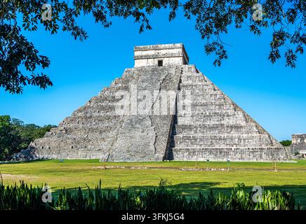 Vista frontale della piramide a gradoni El Castillo, nota anche come Tempio di Kukulkan, nel sito archeologico di Chichen Itza vicino a Mérida nello Yucatan, Messico. L'antica piramide di pietra Maya si erge contro un cielo blu brillante, incorniciata da erba verde e fogliame Foto Stock