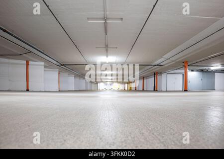 Vista ad angolazione bassa che si concentra sul pavimento in cemento strutturato nell'interno di un parcheggio sotterraneo vuoto con pilastri di sostegno arancioni, pareti bianche e soffitto Foto Stock