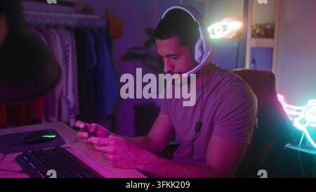 Uomo con auricolari tiene un gamepad e uno smartphone, toccando lo schermo con mani concentrate in uno studio di streaming; concentrazione. Foto Stock