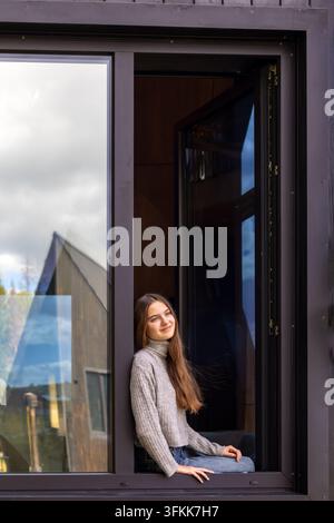 Giovane donna seduta nella finestra aperta di una cabina moderna. Giovane donna seduta nella finestra aperta di una moderna cabina di legno, sorridendo dolcemente alla telecamera. Foto Stock