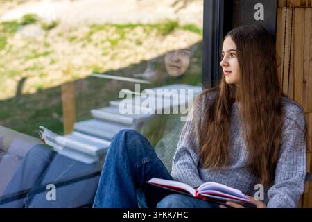Giovane donna che legge vicino a una finestra con riflesso. Giovane donna seduta vicino a una grande finestra tiene un libro aperto e guarda fuori, con il suo riflesso visibile Foto Stock