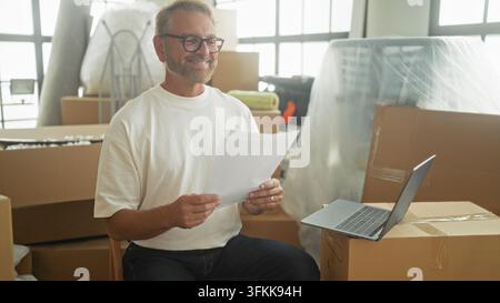 Uomo che legge un giornale accanto a un laptop tra scatole piene in uno studio, fa un pugno in aria mentre sorride; celebrazione di sollievo. Foto Stock