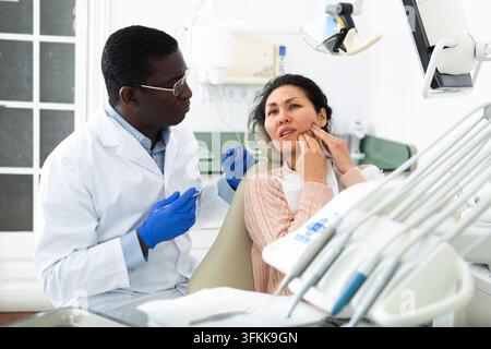 Una paziente che è venuta a un appuntamento con un dentista maschio è seduta sulla poltrona dentale nello studio, mostrando da che lato le fa male il dente Foto Stock
