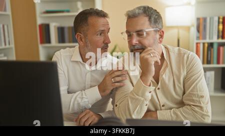 Uomo che sussurra all'orecchio del partner accanto a un laptop e un monitor, mano sul mento e si china in un edificio con scaffali e una lampada; intimità fiducia co Foto Stock