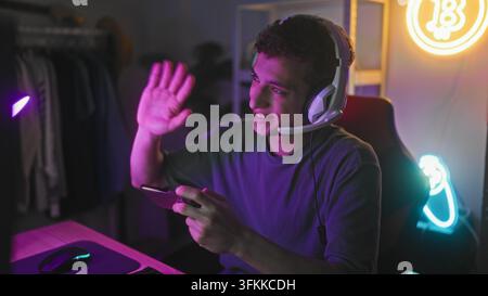 Giovane uomo con auricolari tiene uno smartphone e agita la mano mentre è seduto in un setup di gioco da camera da letto; gioia amichevole nel gaming. Foto Stock
