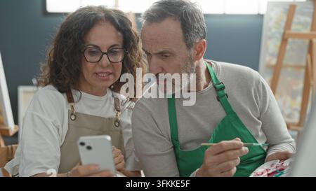 Donna indica lo schermo dello smartphone mentre l'uomo dipinge in studio con un cane piccolo in grembo e cavalletto nelle vicinanze; collaborazione di concentrazione. Foto Stock