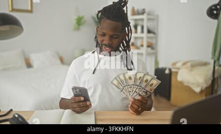 Uomo sventola dollari mentre controlla lo smartphone in camera da letto con notebook e laptop sulla scrivania; celebrazione di gioia e successo. Foto Stock