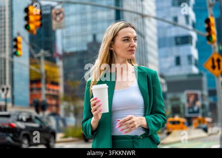 Giovane donna d'affari con capelli biondi e un completo verde che porta una tazza di caffè monouso mentre è in piedi su una strada cittadina con grattacieli e traffico Foto Stock