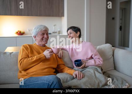 Allegra coppia anziana ride insieme, beve il tè e si rilassa sul divano a casa al chiuso Foto Stock