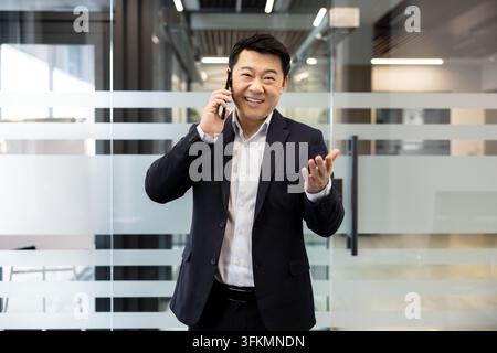 Uomo d'affari che parla al smartphone in un ufficio moderno, uomo professionale in una telefonata. Comunicazione aziendale e discussione commerciale con un manager sorridente che lavora in un luogo di lavoro luminoso. Foto Stock