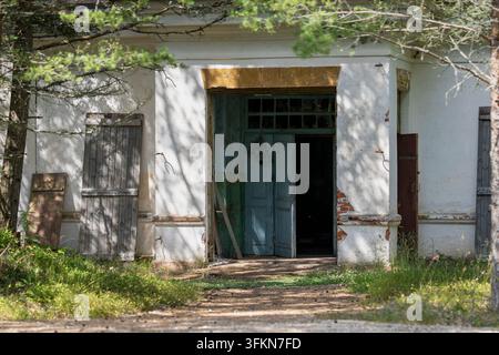 Edificio militare abbandonato a Ristna, in Estonia, con una facciata bianca consumata dal tempo, porte di legno blu-verdi scrostate, finestre sbarrate e muratura a vista. Foto Stock
