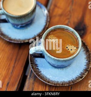 Vista ad angolo alto di due tazze di ceramica artigianali piene di caffè nero caldo con una ricca crema dorata. Servite su piattini abbinati su un ru naturale Foto Stock