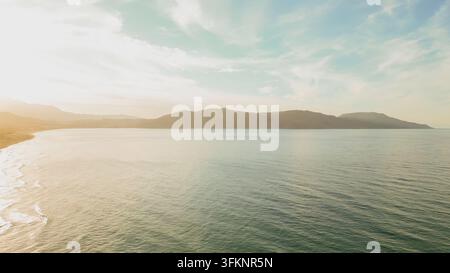 Vista aerea panoramica della costa marina con onde dolci, montagne lontane e luce calda di un tramonto soffuso sull'isola di Creta durante un viaggio estivo in Grecia. Foto Stock