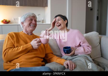 Coppia di anziani felici che beve tè e ride insieme durante una conversazione rilassata a casa sul divano Foto Stock