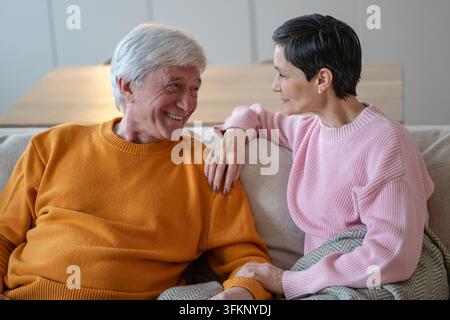 Amorevole moglie anziana tiene la mano del marito mentre la coppia di anziani parla insieme sul divano in casa Foto Stock
