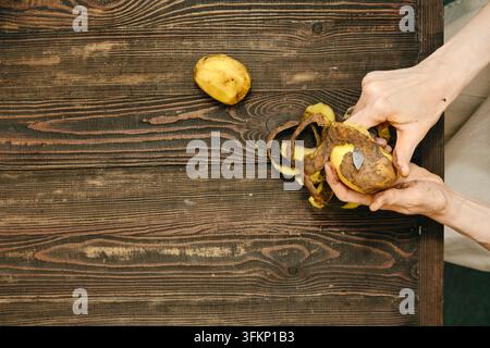 Vista dall'alto di una patata pelata con un coltello su un tavolo di legno in cucina. La buccia della patata viene rimossa in un contesto semplice da cucina Foto Stock
