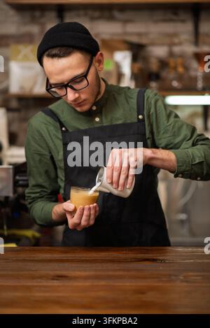 Un barista maschio che versa latte vaporizzato da un contenitore in un bicchiere di caffè Foto Stock