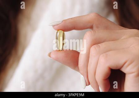 Donna in accappatoio bianco che tiene una capsula di vitamina gialla vicino al viso. Sfondo benessere per integratori nutrizionali quotidiani, cura di sé e stile di vita sano. Foto Stock