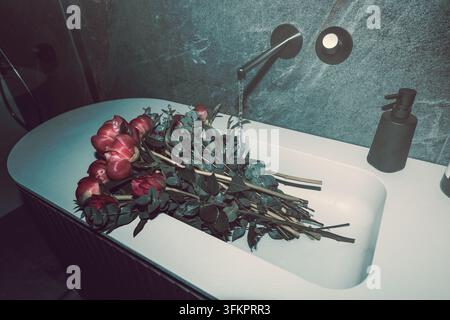 L'acqua scorre da un rubinetto a parete nero su peonie e rose sparse su un lavandino bianco in un bagno contemporaneo di design italiano. Foto Stock