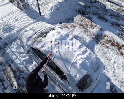 Un uomo pulisce la neve da un'auto in una zona innevata di primo mattino. Il tempo invernale ricopre il veicolo di neve. Foto Stock