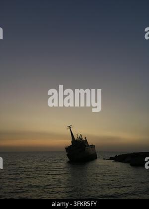 Un relitto di nave si inclina verso la riva a Paphos, Cipro, sullo sfondo di un cielo al tramonto con una sfumatura di blu e arancio. La nave, in silhouette contro il Foto Stock