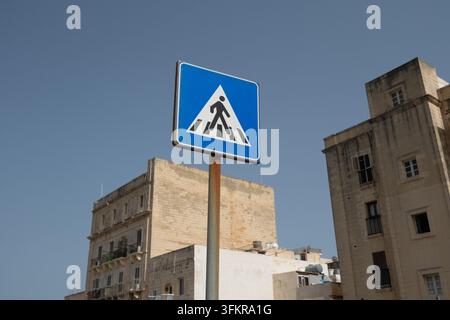 Un cartello stradale blu con un triangolo e una grafica di un omino che attraversa la strada situato a La Valletta, Malta. Foto Stock