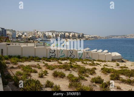 Una vista di St Pauls Bay Malta con un muro bianco in primo piano con graffiti con il tag Elro accanto a terreno incolto, Foto Stock