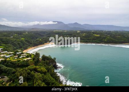 Vista aerea della costa di Kauai con acqua turchese calma, spiaggia dorata curva, fitta foresta tropicale, case sparse e creste montuose stratificate u Foto Stock