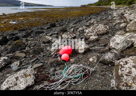 Corda marina arrotolata e boe marcatori luminosi appoggiati tra i massi sulla battigia intertidale, fondo marino esposto con alghe e colline sullo sfondo Foto Stock