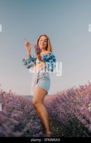 Donna lavanda nel campo che sorride mentre tiene un mazzo di fiori viola in un top floreale e pantaloncini di jeans al tramonto Foto Stock