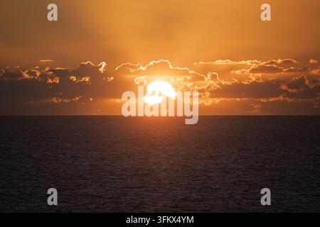Il sole che si fa strada attraverso nuvole retro illuminate sopra l'orizzonte del mare con raggi in un cielo arancione dorato. Foto Stock