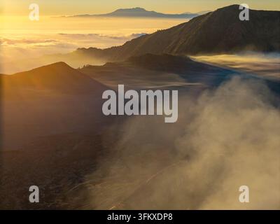 L'alba dorata avvolge le montagne nebbiose in una luce calda. Ampie nuvole fluttuano sotto le vette vulcaniche frastagliate vicino a Bromo Foto Stock