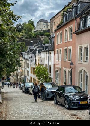 Due donne camminano lungo una storica strada acciottolata fiancheggiata da vecchi edifici colorati, auto parcheggiate e alberi verdi rigogliosi sotto un cielo nuvoloso. Foto Stock