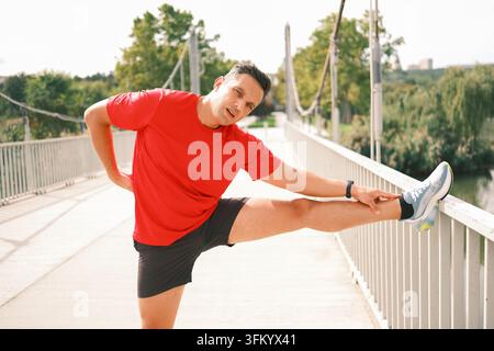 Uomo atletico che allunga la gamba su una ringhiera del ponte prima o dopo la corsa, migliorando la flessibilità e la mobilità durante un allenamento di fitness all'aperto Foto Stock