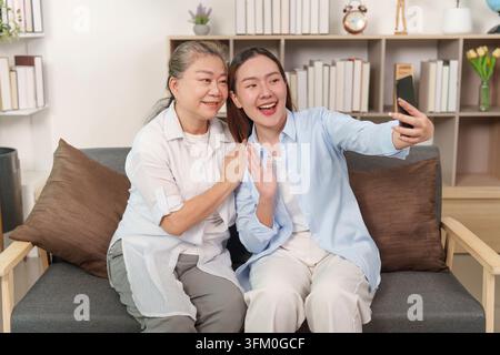 Una figlia asiatica e sua madre anziana si impegnano felicemente in una videochiamata usando uno smartphone mentre sono sedute su un divano a casa. Questa immagine raffigura moderna i Foto Stock