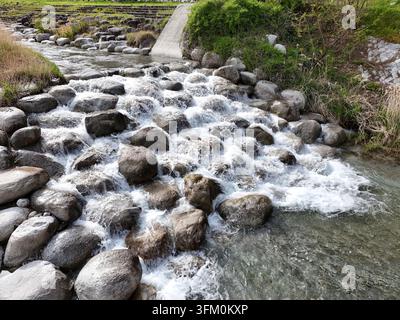 L'acqua scorre su pietre grigie lisce. La schiuma bianca balla mentre il flusso scorre verso il basso. L'erba verde cresce accanto al letto del fiume roccioso. Vento su percorso in cemento Foto Stock