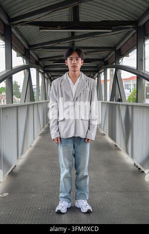 Autentico ritratto verticale a tutta lunghezza di un giovane professionista isano thailandese non binario o di uno studente in piedi simmetricamente su un ponte pedonale. Guardando Foto Stock