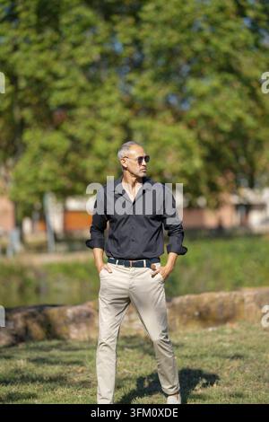Un uomo con una camicia nera e pantaloni abbronzati si trova in un campo erboso. Indossa gli occhiali da sole e ha le mani nelle tasche. Concetto di relax e le Foto Stock