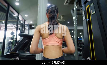 Una vista posteriore di una giovane donna asiatica con una coda di cavallo che indossa un reggiseno sportivo pesca e leggings neri, in piedi in una moderna palestra con tapis roulant Foto Stock
